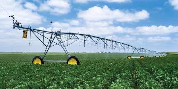 A large center pivot irrigation system spans across a green field, spraying water over crops under a clear blue sky.